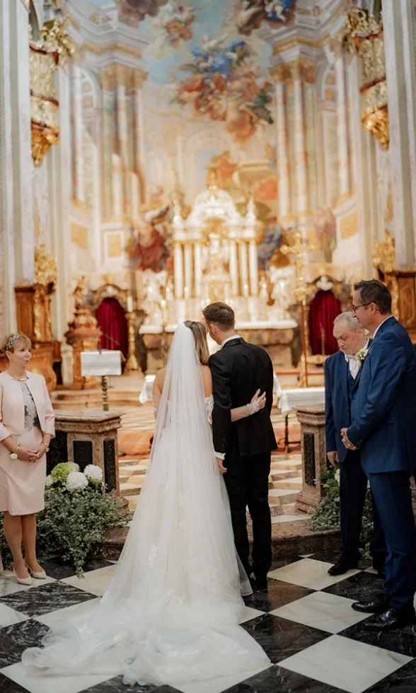 Braut und Bräutigam stehen vor dem Altar in der prunkvollen Kirche, umgeben von Familie während der Trauung.