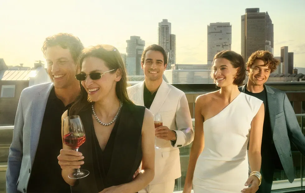 Gruppe junger Menschen in eleganter Digel-Mode auf einer sonnigen Rooftop-Party, Männer in modernen Anzügen und Frauen in stylishen Kleidern, lässig mit Drinks in der Hand.