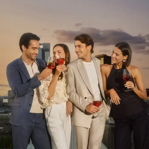 Freundesgruppe in Digel-Anzügen und modernen Outfits, die bei Sonnenuntergang auf einer Rooftop-Bar anstoßen; stilvolle Abendstimmung und elegante Menswear.