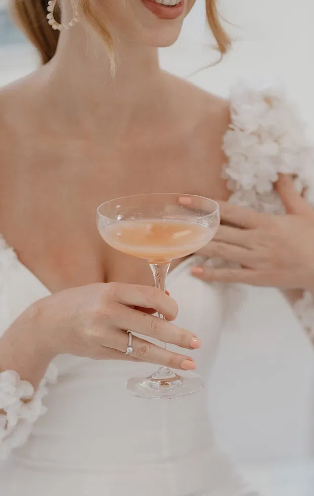 Braut im weißen Kleid hält einen zarten pfirsichfarbenen Mocktail in einer Coupette beim Getting Ready.