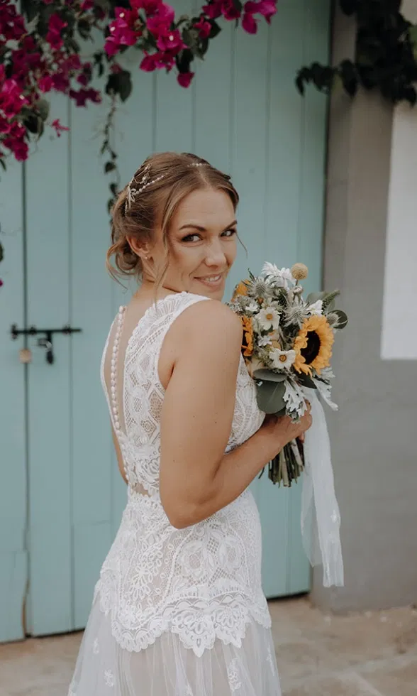 Lächelnde Braut im Spitzenkleid blickt über die Schulter und hält einen Strauß mit Sonnenblumen vor mintfarbener Tür.