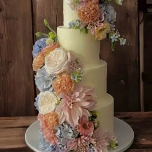Mehrstöckige Hochzeitstorte in zartem Cremeweiß, dekoriert mit üppigen floralen Arrangements aus pastellfarbenen Blüten wie Dahlien, Rosen und Hortensien, vor rustikalem Holz­hintergrund.