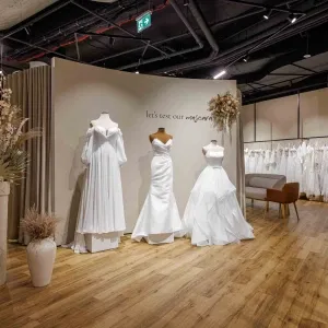 Innenansicht eines modernen Brautmodengeschäfts mit drei ausgestellten Brautkleidern auf Schaufensterpuppen, warmem Holzfußboden, neutralem Farbkonzept und weiteren Brautkleidern im Hintergrund.