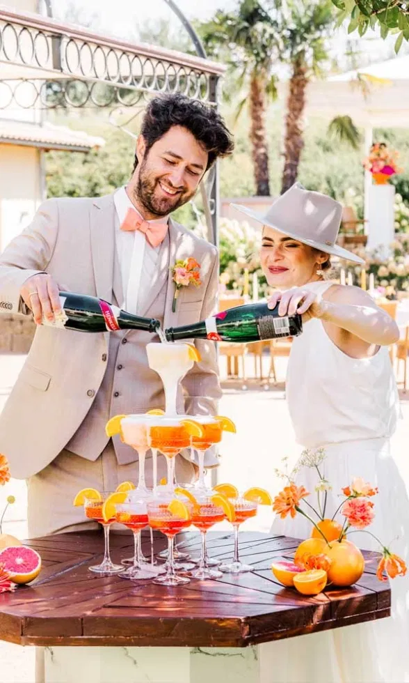 Ein fröhliches Brautpaar schenkt gemeinsam Sekt in einen pyramidenförmig aufgebauten Aperitif-Tower mit orangefarbenen Drinks und frischen Zitrusscheiben, vor einer sonnigen Outdoor-Hochzeitskulisse.