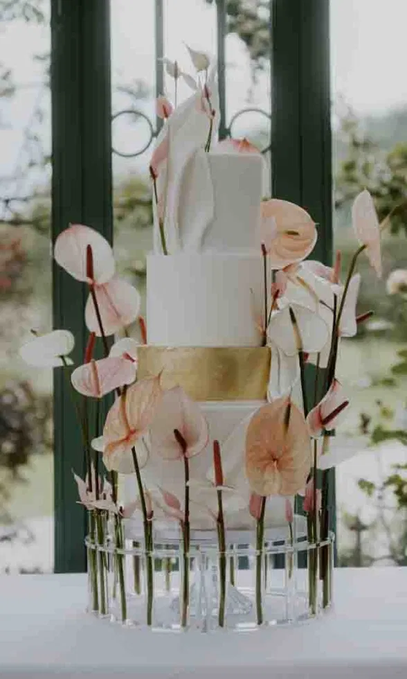 Moderne Hochzeitstorte mit klaren Linien, weißem Fondant, goldenem Akzentband und zarten Anthurienblüten in Glasröhrchen arrangiert.
