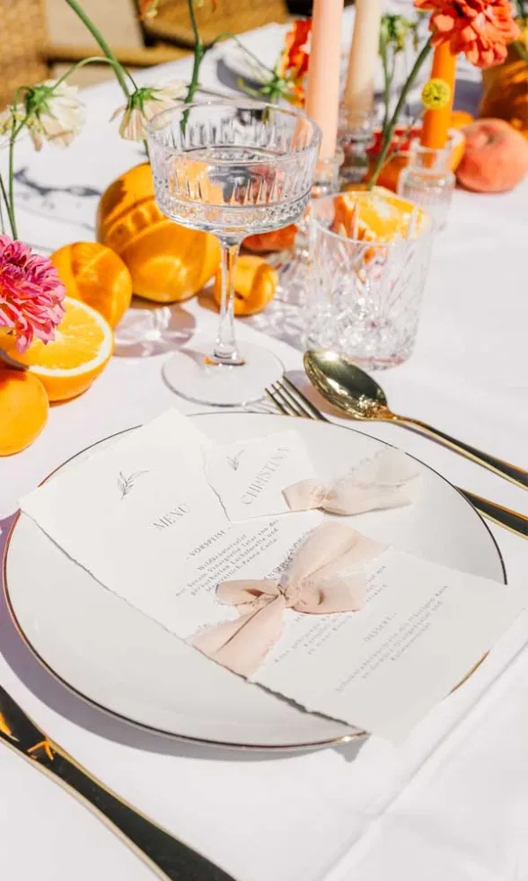 Detailaufnahme einer sommerlich gedeckten Hochzeitstafel mit Coupette-Glas, Kristallbecher, goldfarbenem Besteck und handgerissener Menükarte auf dem Teller, ergänzt durch Orangen, florale Akzente und warmes Sonnenlicht.