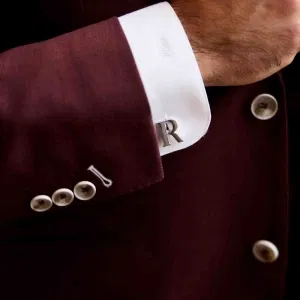 Detailaufnahme eines Bräutigams mit bordeauxfarbenem Anzug und Manschettenknopf mit Initiale am weißen Hemd.