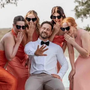 Bräutigam sitzt lachend im Mittelpunkt, umgeben von Brautjungfern in warmen Farbtönen, die spielerisch reagieren.