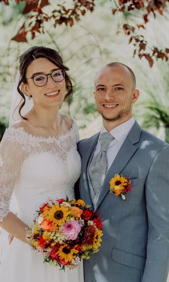 Glückliches Brautpaar steht im Grünen und lächelt in die Kamera, die Braut im Spitzenbrautkleid mit Brille und buntem Blumenstrauß, der Bräutigam im hellblauen Anzug mit Ansteckblume.