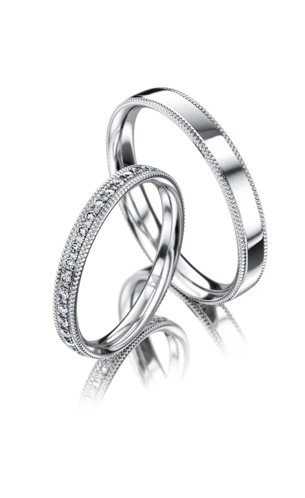 Zwei filigrane Eheringe aus Weißgold mit feiner Millegriffes-Verzierung, einer davon mit einer schmalen Reihe eingefasster Diamanten, elegant im Vintage-Stil vor hellem Hintergrund fotografiert.