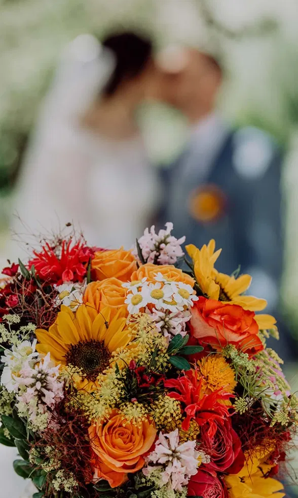 Farbenfroher Brautstrauß mit Sonnenblumen, Rosen und Wildblumen im Vordergrund, im Hintergrund unscharf das Brautpaar beim Kuss.