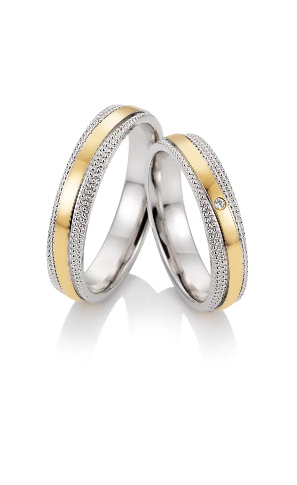 Zwei Eheringe im Bicolor-Design aus Weiß- und Gelbgold mit fein strukturierter Oberfläche, einer davon mit dezent gefasstem Diamanten, klassisch im Vintage-Stil vor hellem Hintergrund fotografiert.