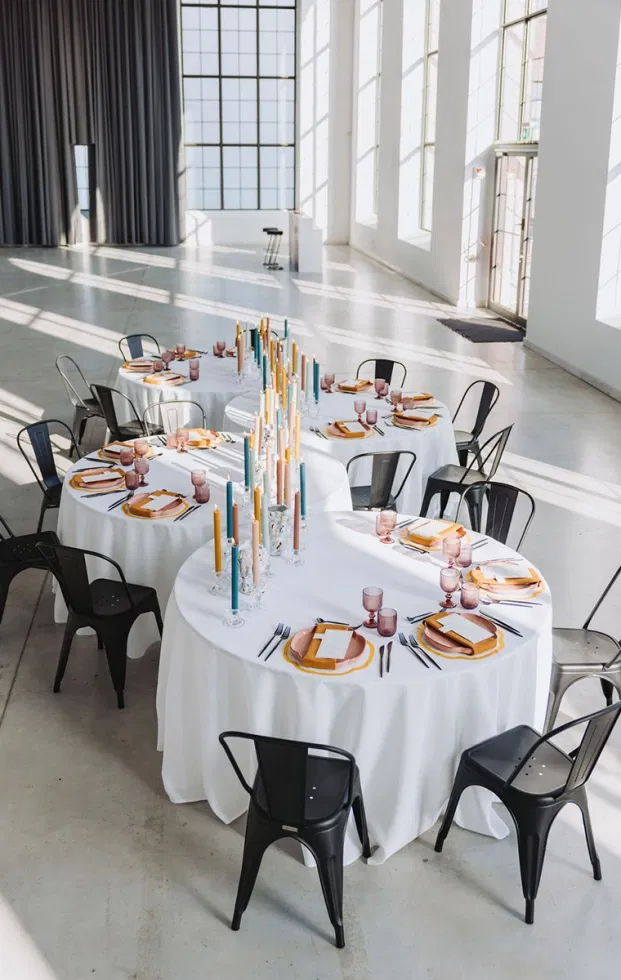 Großzügige Hochzeitslocation mit runden Tischen, Artsy Wedding Table Settings, bunten Kerzen und moderner Industrie-Ästhetik.