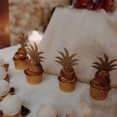 Kleine Cupcakes in Goldtönen mit cremigem Topping und dekorativen Ananas-Toppern, arrangiert auf einem stilvollen Sweet Table bei Kerzenschein.