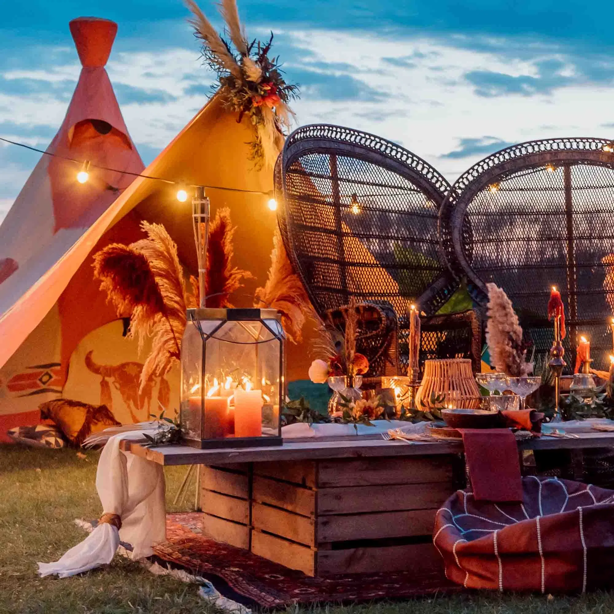 Stimmungsvoll dekorierter Tisch im Festival-Chic mit Kerzen, Pampasgras und warmem Licht vor einem Tipi-Zelt bei Sonnenuntergang.