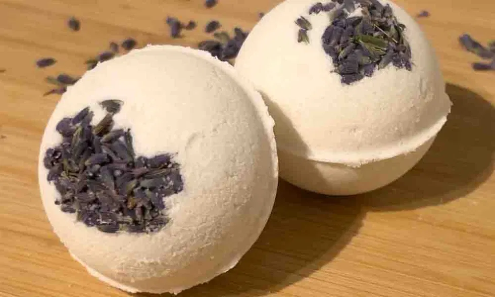 Zwei selbstgemachte Badekugeln mit getrockneten Lavendelblüten liegen auf einer Holzoberfläche.