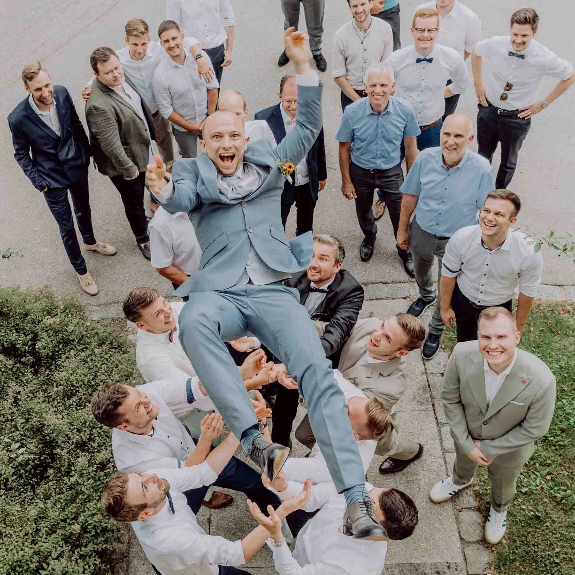 Bräutigam im hellblauen Anzug wird lachend von seinen Freunden in die Luft geworfen, umgeben von Hochzeitsgästen bei ausgelassener Stimmung im Freien.