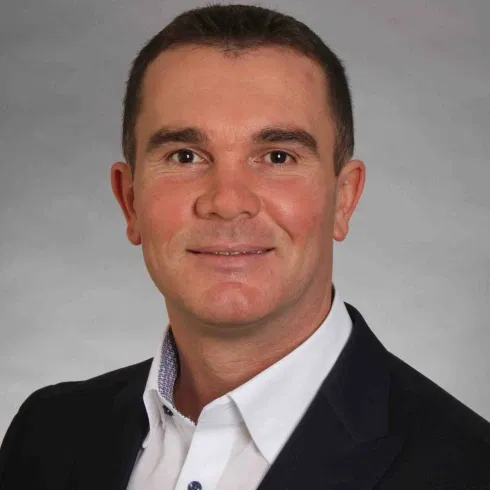 Porträt eines Mannes im dunklen Sakko und weißen Hemd vor neutralem, grauem Hintergrund.