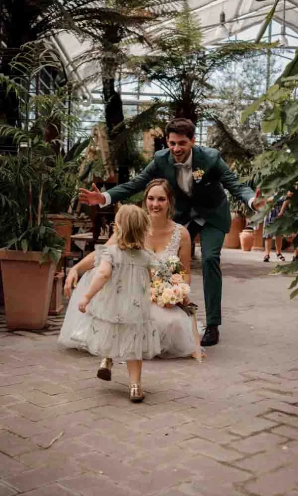 Rührender Hochzeitsmoment im Gewächshaus: Das Brautpaar kniet lächelnd, während ihr kleines Kind auf sie zuläuft, umgeben von üppigem Grün
