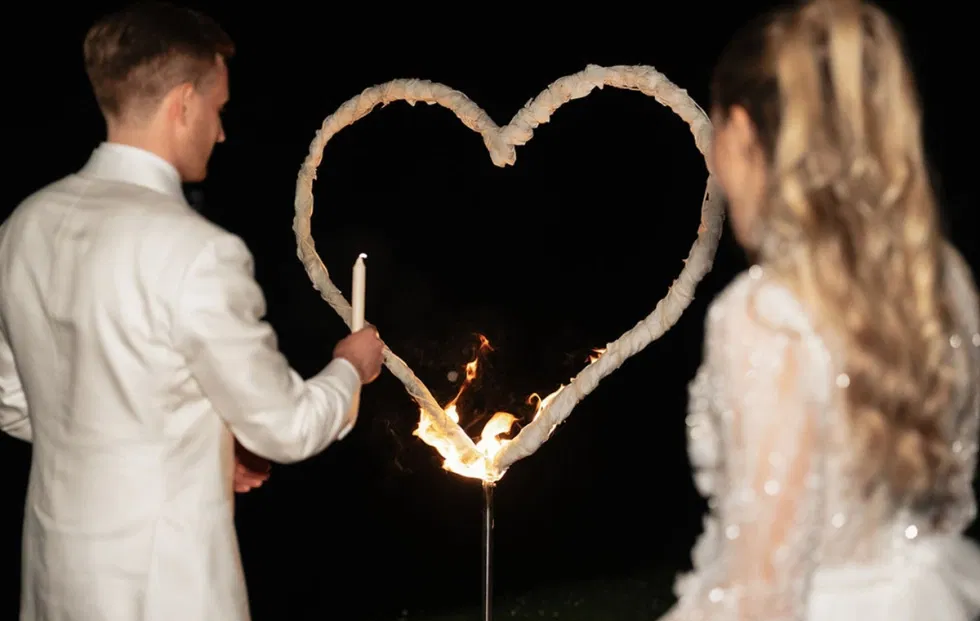 Braut und Bräutigam entzünden gemeinsam ein brennendes Herz aus Flammen als symbolischen Hochzeitsabschluss in stimmungsvoller Abendatmosphäre.