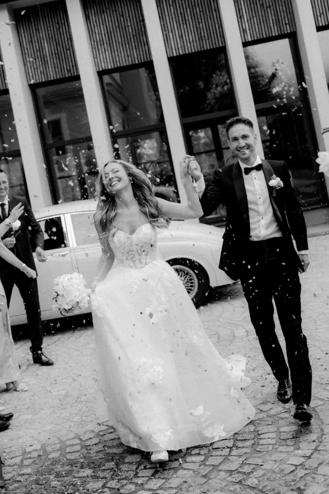 Schwarz-weiß Aufnahme des Brautpaares beim Auszug nach der Trauung, Hand in Hand lachend, während Blütenblätter durch die Luft fliegen, die Braut im floralen Brautkleid und der Bräutigam im eleganten Smoking vor klassischer Architektur und Oldtimer.