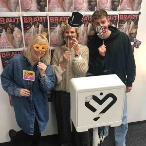 Drei Personen stehen lachend vor einer Fotobox und halten lustige Foto-Requisiten wie Masken, Schnurrbart und Schriftzüge in den Händen. Im Hintergrund sind mehrere Cover des Hochzeitsmagazins „Braut“ an der Wand befestigt.