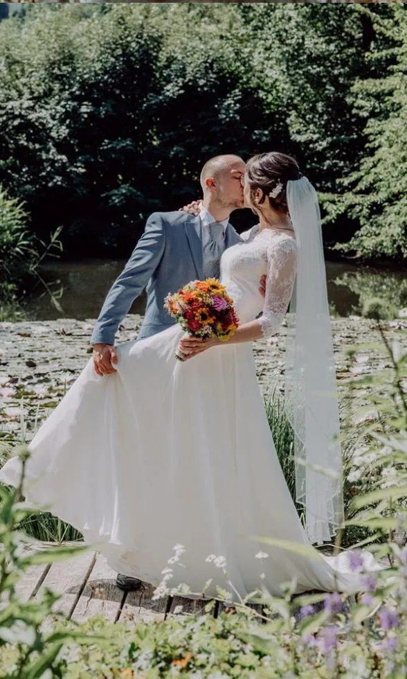 Braut und Bräutigam küssen sich romantisch auf einem Holzsteg am See, umgeben von üppigem Grün, die Braut im fließenden Kleid mit Schleier und buntem Brautstrauß, ein inniger Moment in natürlicher Kulisse.