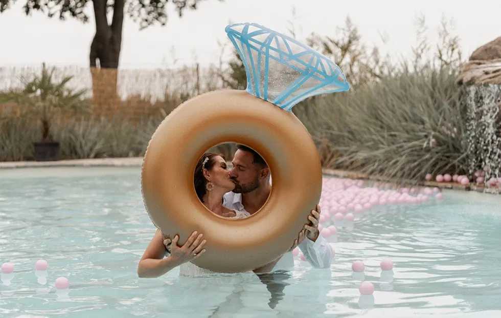 Brautpaar küsst sich verspielt im Pool durch einen Schwimmring mit Diamantmotiv – sommerliche Pre-Wedding-Week-Stimmung vor der Hochzeit.
