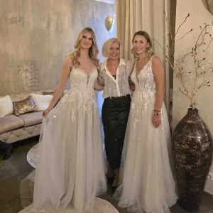 Drei Frauen stehen lächelnd in einem stilvollen Bridal-Showroom: Zwei Bräute in fließenden, hellen Brautkleidern mit feiner Spitze flankieren eine ältere Frau in eleganter Bluse und dunklem Rock, umgeben von warmer, wohnlicher Atmosphäre.
