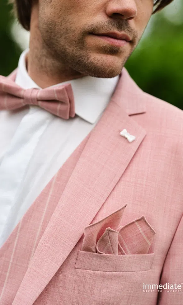 Detailaufnahme eines Bräutigams im roséfarbenen Anzug mit Fliege, Einstecktuch und kleiner Reversnadel.