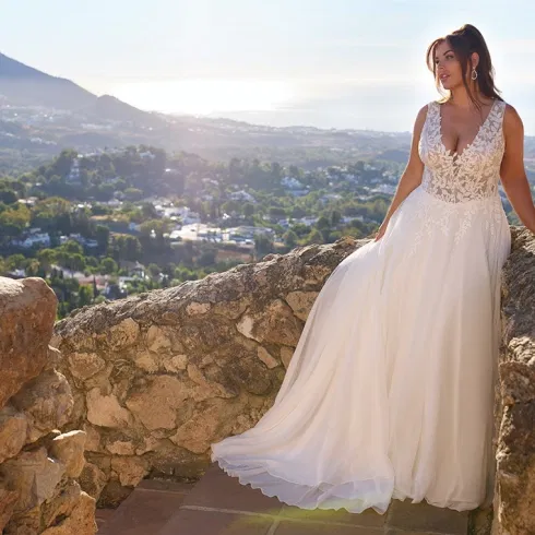 Braut im fließenden A-Linien-Brautkleid mit Spitzenoberteil, fotografiert auf einer Natursteinmauer mit mediterraner Landschaft im Hintergrund.