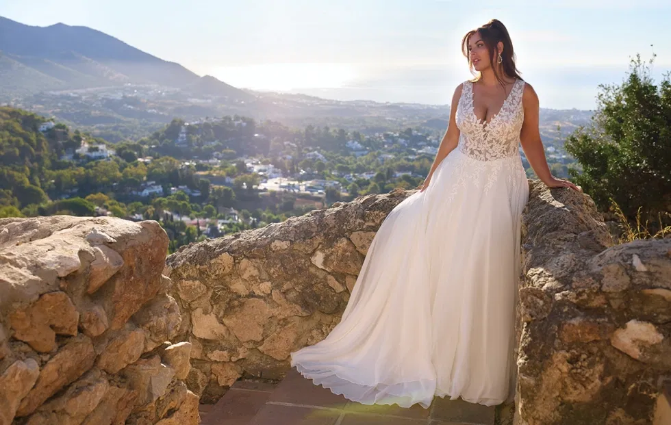 Braut im fließenden A-Linien-Brautkleid mit Spitzenoberteil, fotografiert auf einer Natursteinmauer mit mediterraner Landschaft im Hintergrund.