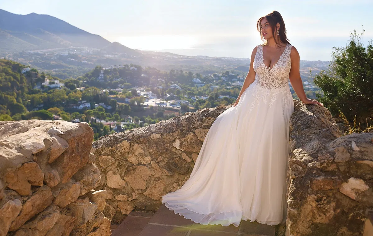 Braut im fließenden A-Linien-Brautkleid mit Spitzenoberteil, fotografiert auf einer Natursteinmauer mit mediterraner Landschaft im Hintergrund.