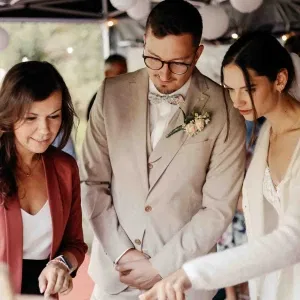 Brautpaar steht gemeinsam mit einer Hochzeitsplanerin an einem Tisch, betrachtet aufmerksam Details der Feier, der Bräutigam im beigen Anzug mit Fliege, die Braut in hellem Outfit, umgeben von warmer, festlicher Atmosphäre.