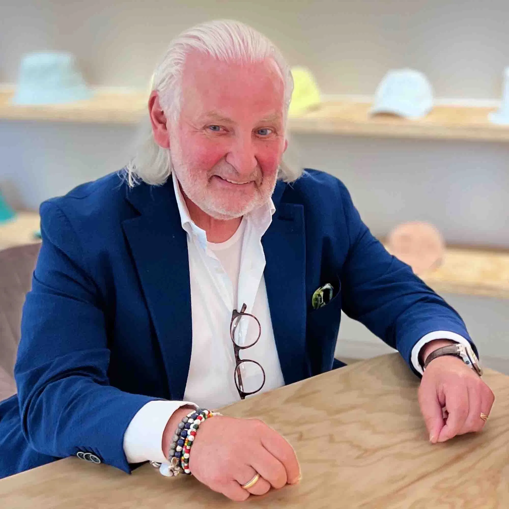 Älterer Mann mit weißem Haar und Bart sitzt an einem Holztisch, trägt ein blaues Sakko über weißem Hemd und lächelt freundlich in die Kamera, im Hintergrund stehen dezent präsentierte Hüte in einem hellen Raum.