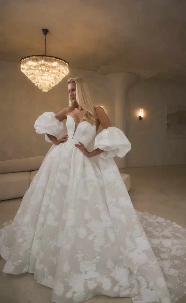 Braut in einem opulenten Prinzessinnen-Brautkleid mit tiefem Herzausschnitt und voluminösen Puffärmeln, inszeniert in einem eleganten Raum mit großem Kronleuchter und fließender Schleppe.