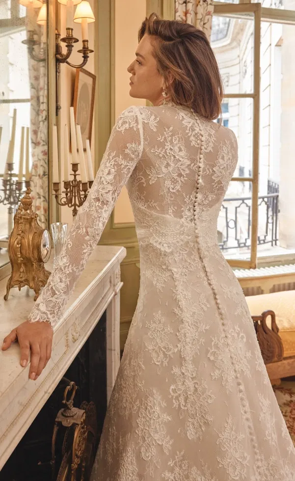Braut in langärmligem Spitzen-Brautkleid mit transparenter Rückenpartie und feiner Knopfleiste, aufgenommen in einem eleganten Salon vor einem Kamin mit Kerzenleuchtern und großem Fenster.