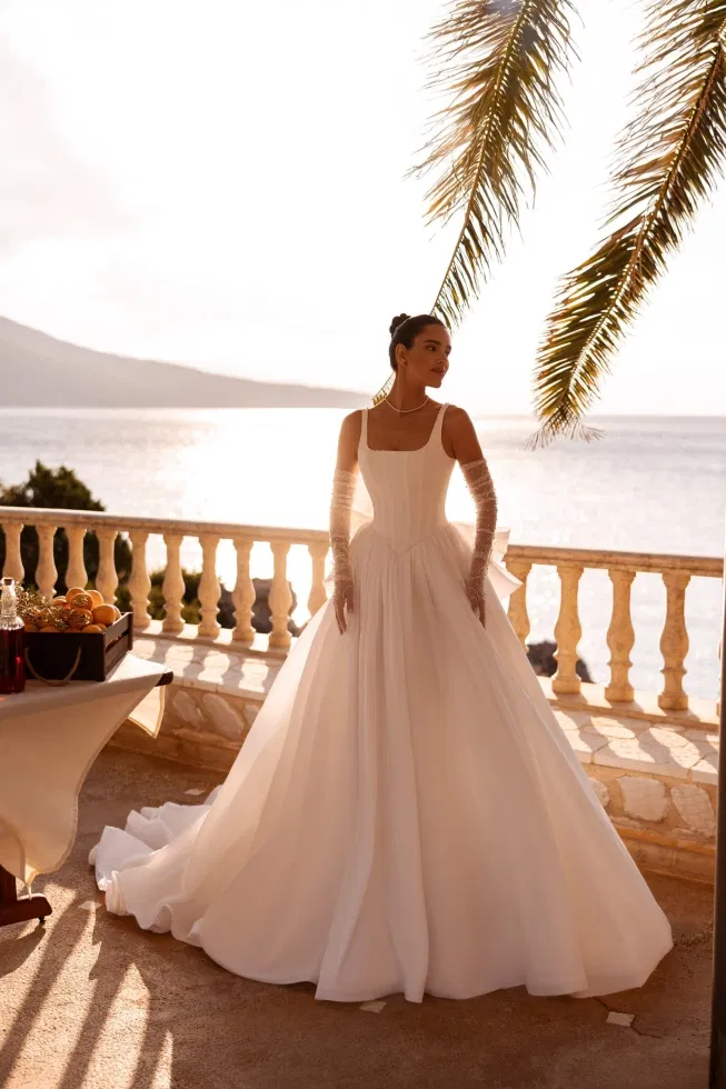 Romantisches Prinzessinnen-Brautkleid mit strukturiertem Korsagenoberteil, langen transparenten Handschuhärmeln und voluminösem Rock, aufgenommen im goldenen Sonnenuntergang am Meer.