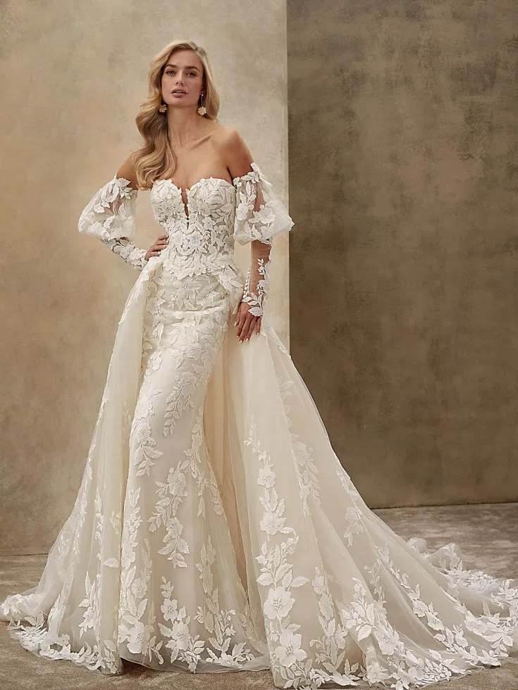 Romantisches Off-Shoulder-Brautkleid in Fit-and-Flare-Silhouette mit floraler 3D-Spitze, transparenten Ärmeln und abnehmbarer Tüllschleppe vor neutralem Studiohintergrund.