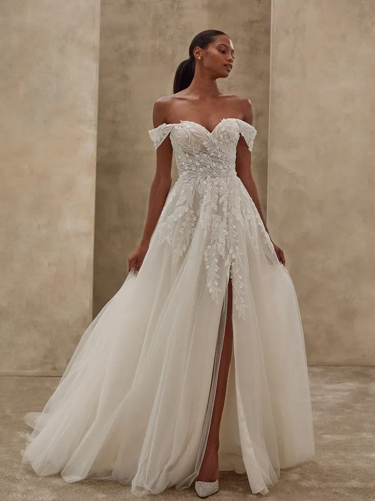 Luftiges A-Linien-Brautkleid mit zarten Off-Shoulder-Trägern, floralen Stickereien und funkelndem Tüllrock mit Schlitz, elegant präsentiert im soften Studioambiente.