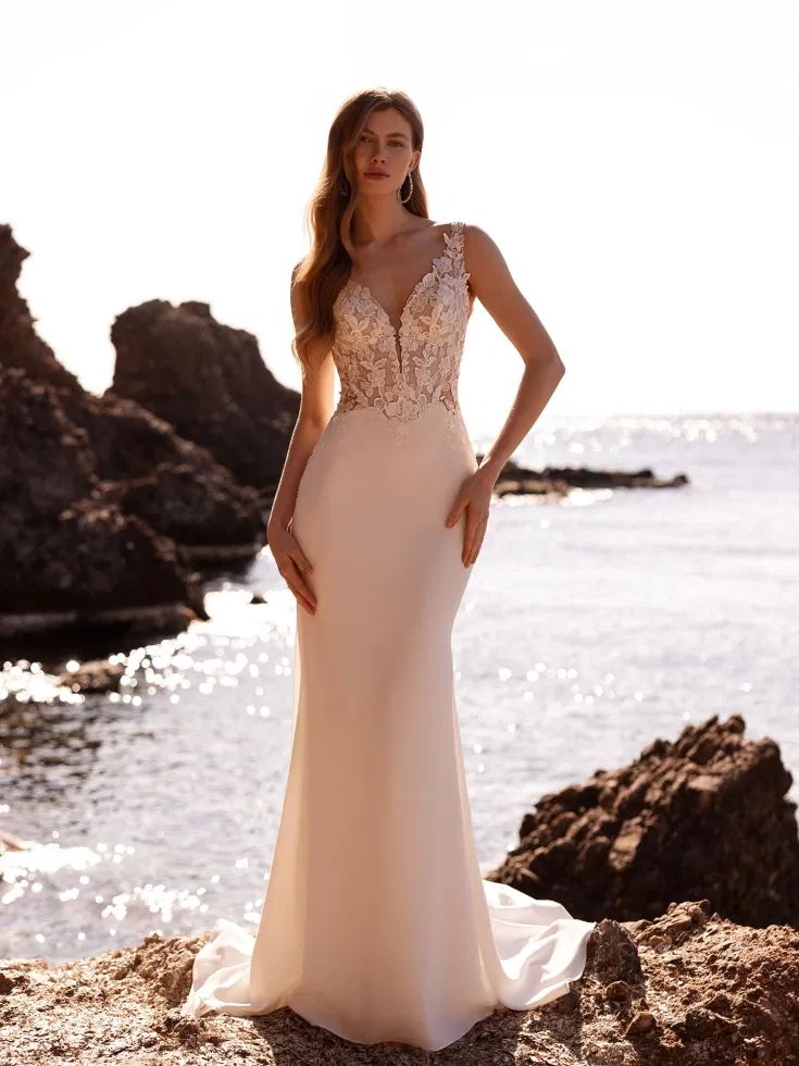 Schlichtes Fit-and-Flare-Brautkleid mit V-Ausschnitt und floraler Spitzenapplikation am Oberteil, fotografiert an einer felsigen Küstenlandschaft im Sonnenlicht.