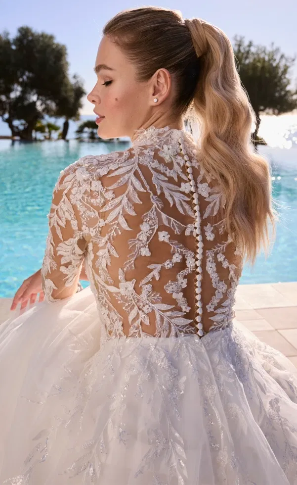 Rückansicht einer Braut mit hochgeschlossenem, transparentem Spitzenoberteil, filigranen floralen Stickereien und eleganter Knopfleiste, vor einem Pool im warmen Sonnenlicht.