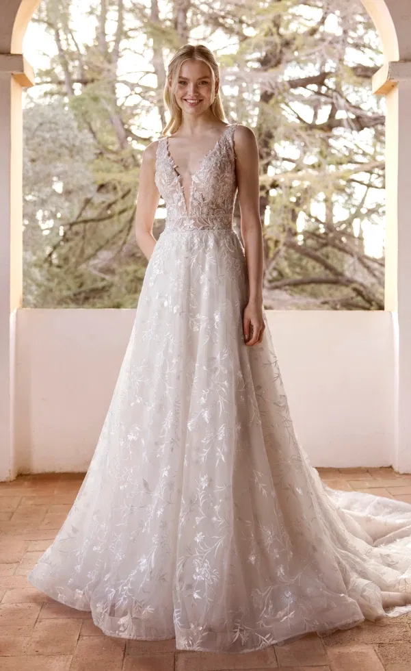 Braut in einem romantischen A-Linien-Brautkleid mit tiefem V-Ausschnitt, zartem Spitzenoberteil und fließendem Rock mit floralen Stickereien, aufgenommen unter einem Torbogen mit Blick ins Grüne.