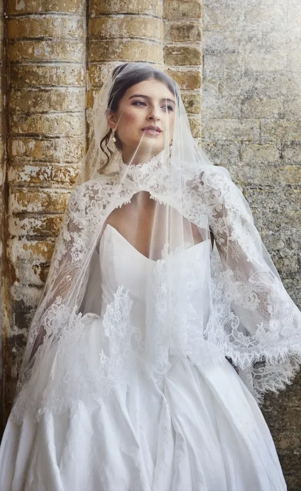 Braut mit klassischem A-Linien-Brautkleid, herzförmigem Ausschnitt und zarter Spitzen-Cape mit Schleier, posierend vor einer historischen Backsteinwand.