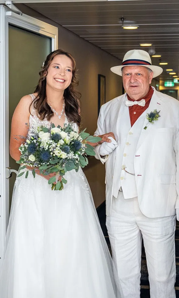Braut mit Brautstrauß wird von einer älteren Begleitperson im hellen Anzug zum First Look geführt, berührender Moment kurz vor der Trauung.