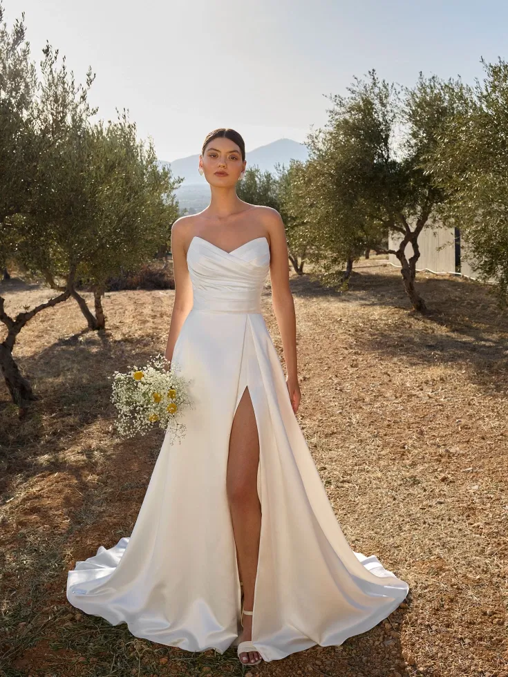 Minimalistisches trägerloses Brautkleid mit drapierter Corsage und hohem Beinschlitz, kombiniert mit einem zarten Brautstrauß in mediterraner Olivenhain-Kulisse.