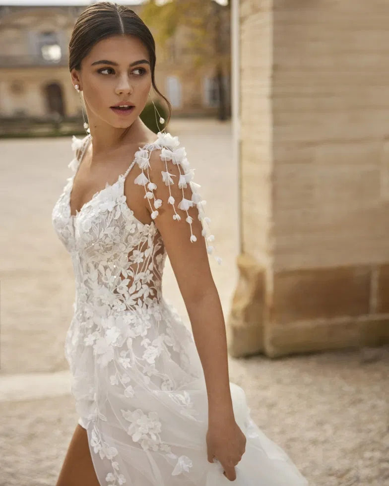 Detailaufnahme einer Braut in einem romantischen Spitzen-Brautkleid mit floralen 3D-Applikationen und zarten, blütenverzierten Schulterträgern vor historischer Architektur.