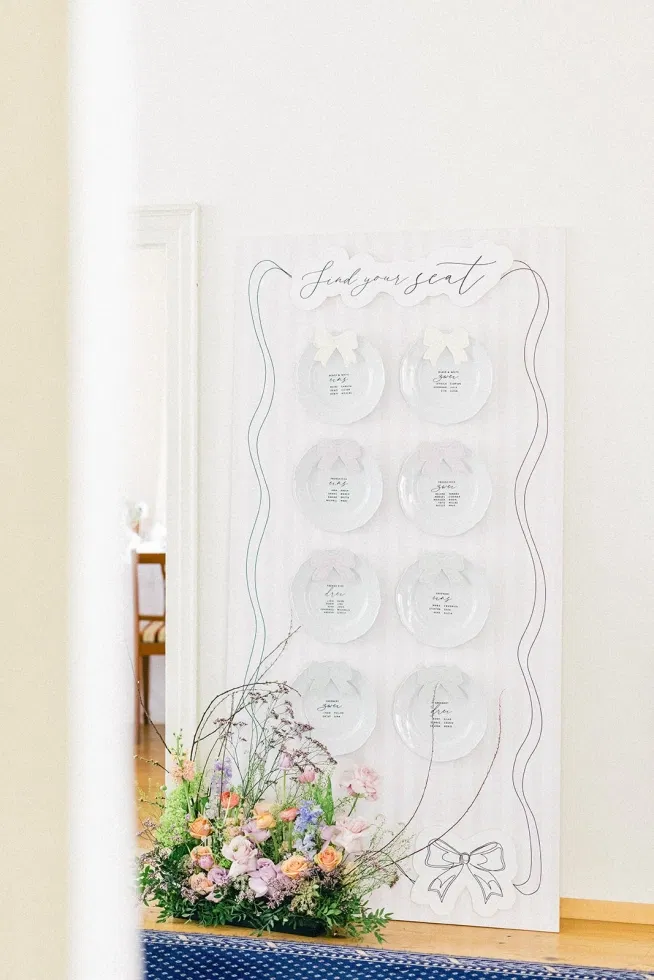 Eleganter Sitzplan für eine Hochzeit mit transparenten Tellern, handgeschriebener Papeterie und floraler Dekoration in zarten Pastellfarben im Fine-Art-Stil.