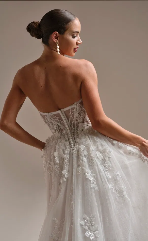 Rückenansicht einer Braut in trägerlosem A-Linien-Brautkleid mit transparentem Korsagenoberteil, zarten Knopfleisten-Details und floralen 3D-Spitzenapplikationen auf dem weich fallenden Tüllrock.