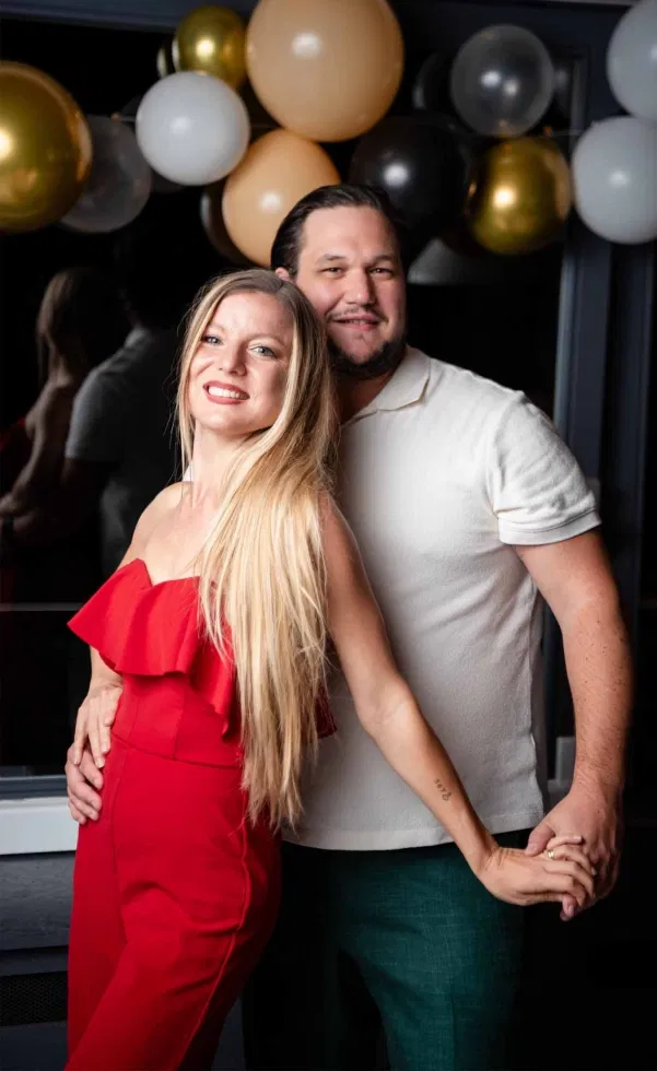 Glückliches Paar vor einer Ballon-Dekoration in Gold-, Schwarz- und Beigetönen: Die Frau trägt einen roten Jumpsuit, der Mann ein helles Shirt mit dunkler Hose – entspannte, persönliche Momentaufnahme mit moderner Feier-Atmosphäre.